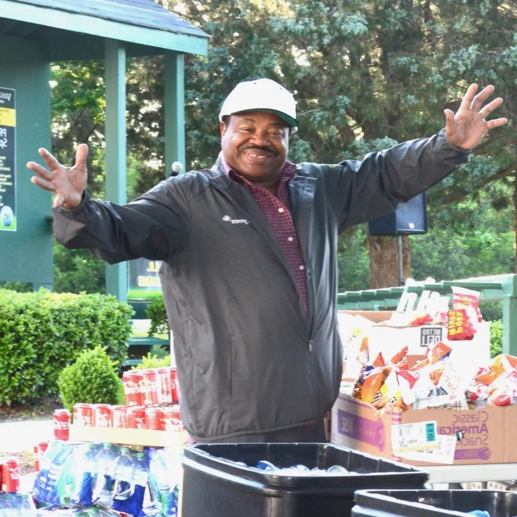 A smiling man holds his arms wide at an outdoor event; he is surrounded by crates of drinks and prepackaged snacks.